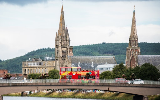 Red double-decker bus on bridge in Inverness with church spires in background.