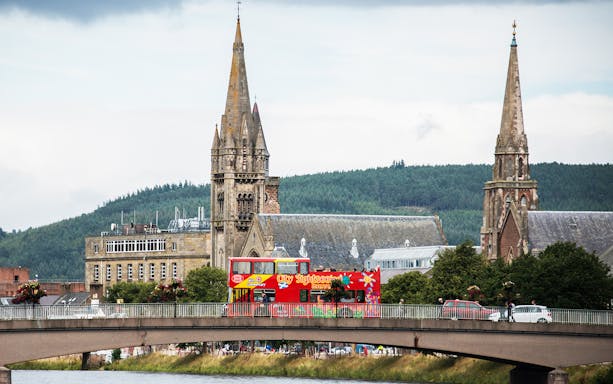 Red double-decker bus on bridge in Inverness with church spires in background.