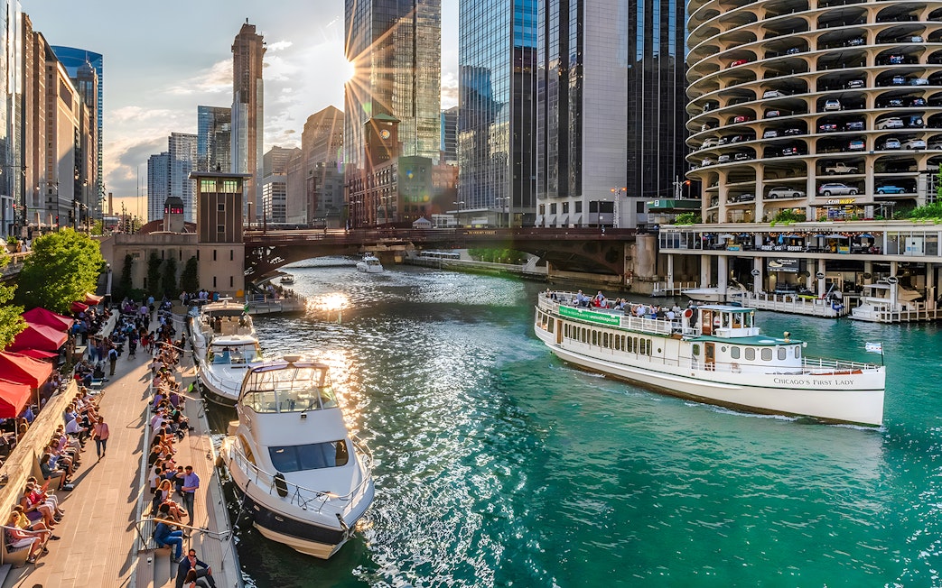 Chicago Riverwalk with people strolling, boats on the river, and 'Chicago's First Lady' tour boat.