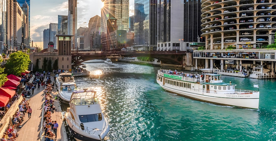 Chicago Riverwalk with people strolling, boats on the river, and 'Chicago's First Lady' tour boat.