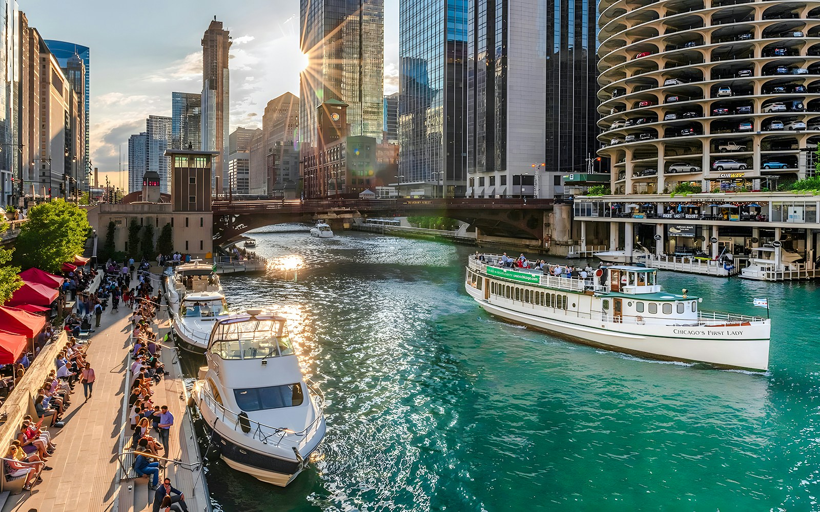 Chicago Riverwalk with people strolling, boats on the river, and 'Chicago's First Lady' tour boat.