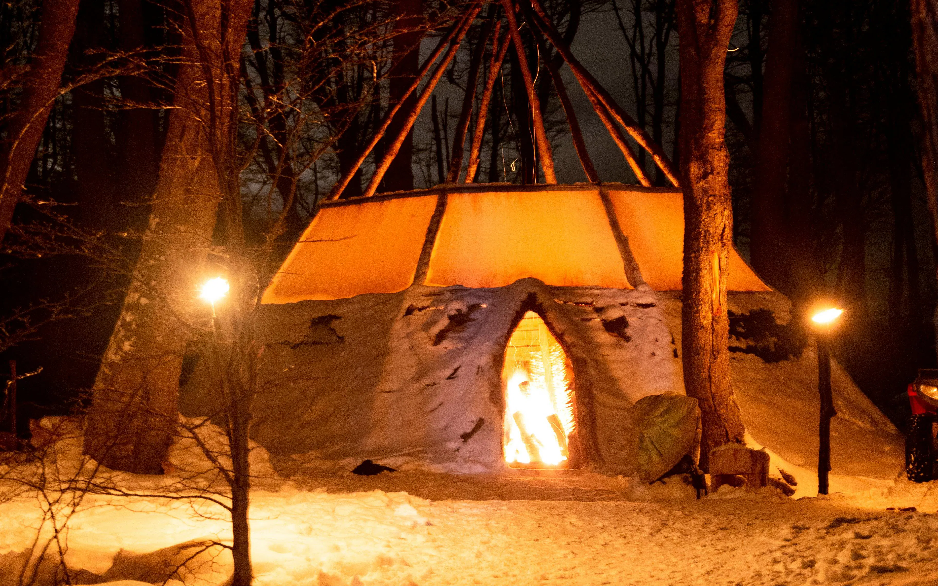 Snow-covered teepee with firelight in a forest during the Snow and Fire Night experience.