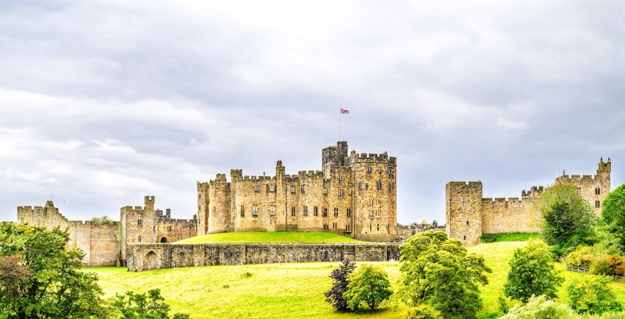 Alnwick Castle with lush green grounds under a cloudy sky in Northumberland, England.