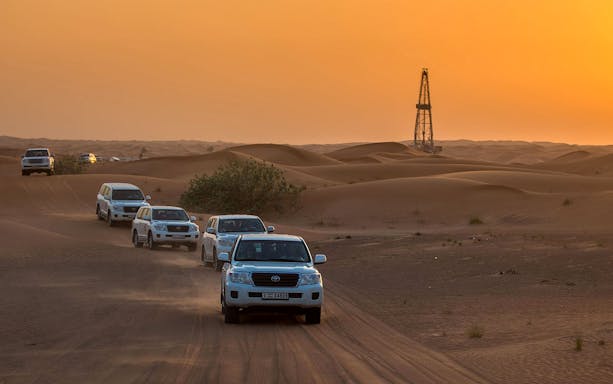 Convoy of SUVs driving through desert dunes at sunset during a premium evening safari.