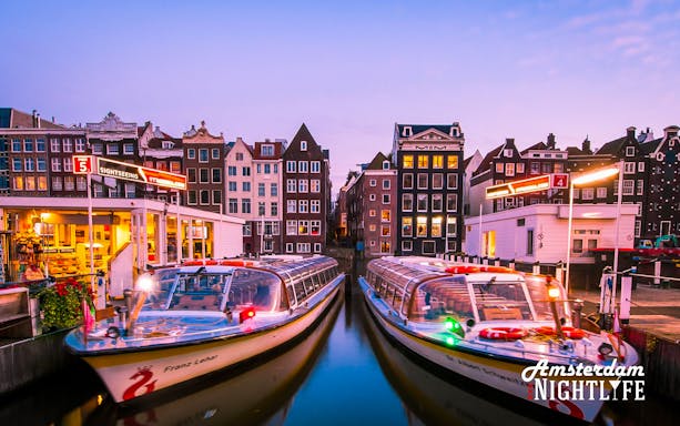 Canal cruise boats docked in Amsterdam at sunset, with historic buildings in the background.