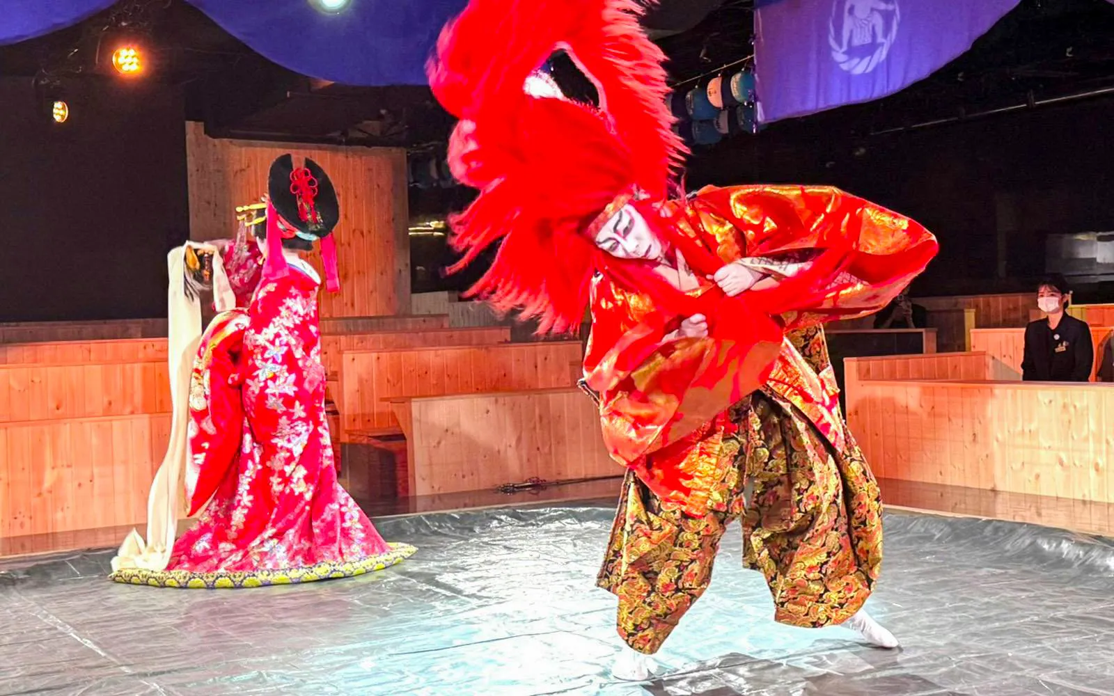 Performer in red lion dance costume with another in ornate attire during Japanese cultural show.