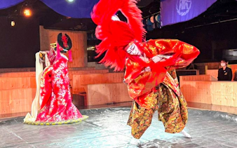 Performer in red lion dance costume with another in ornate attire during Japanese cultural show.