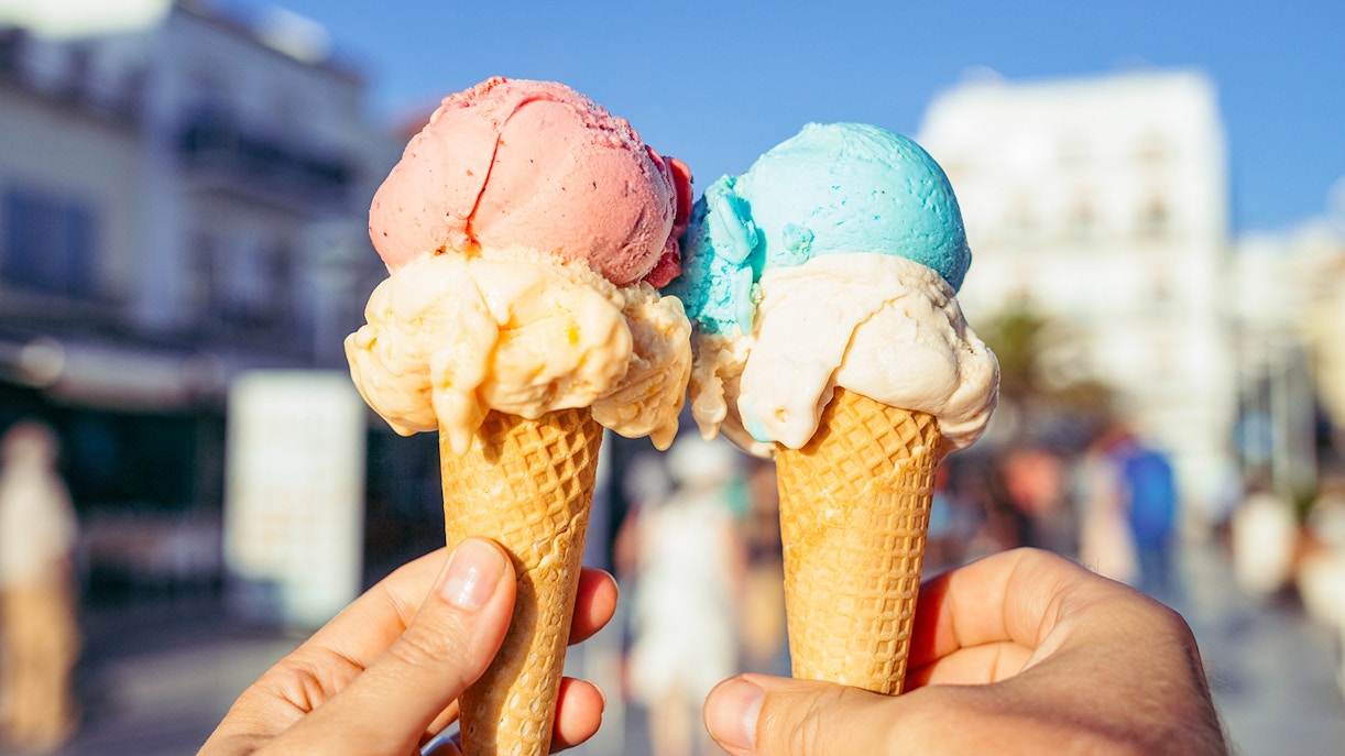 Hands holding waffle cones with pink and blue ice cream in a sunny street.