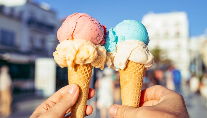 Hands holding waffle cones with pink and blue ice cream in a sunny street.
