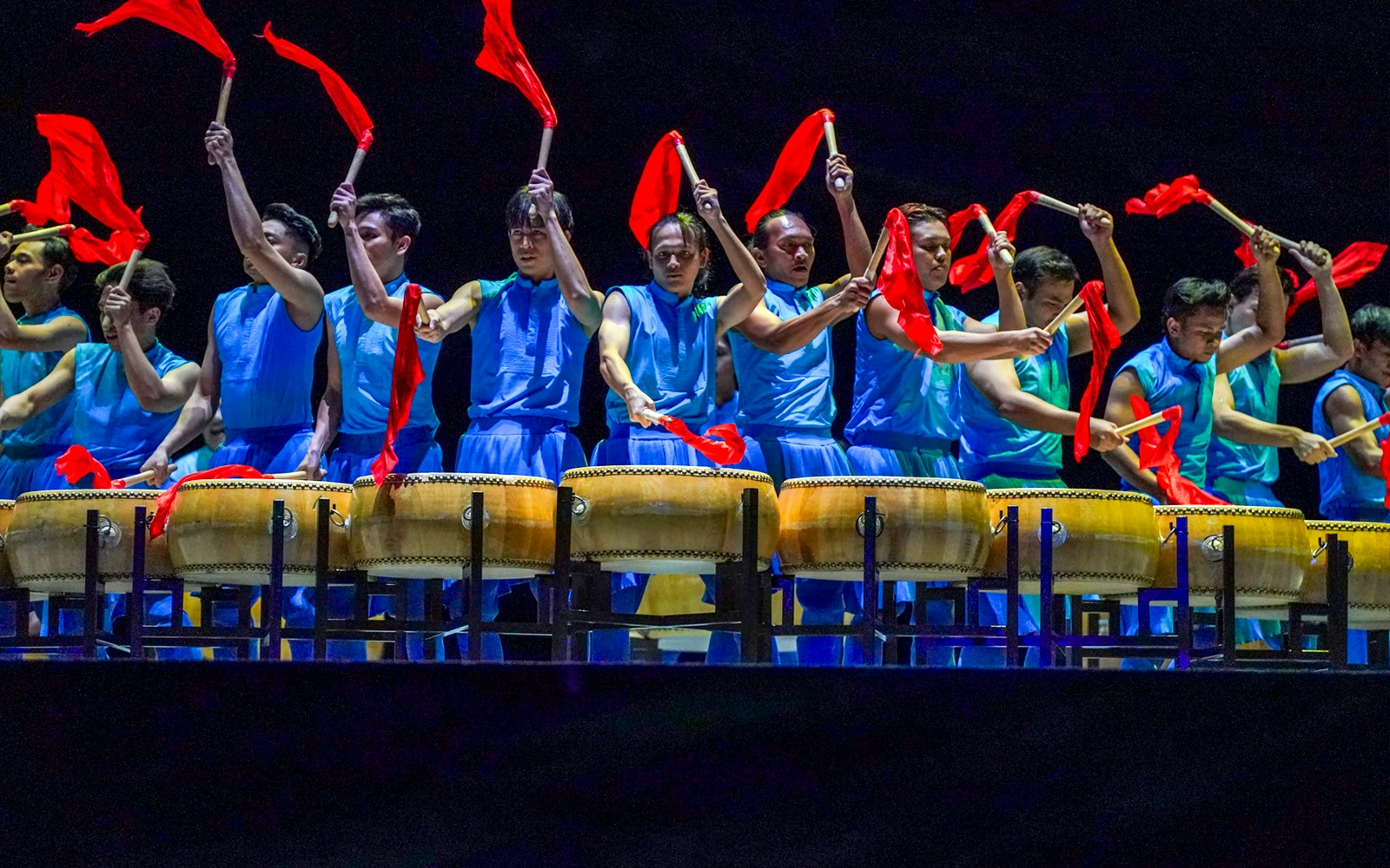 Performers in blue costumes playing drums with red flags at Encore Melaka show.