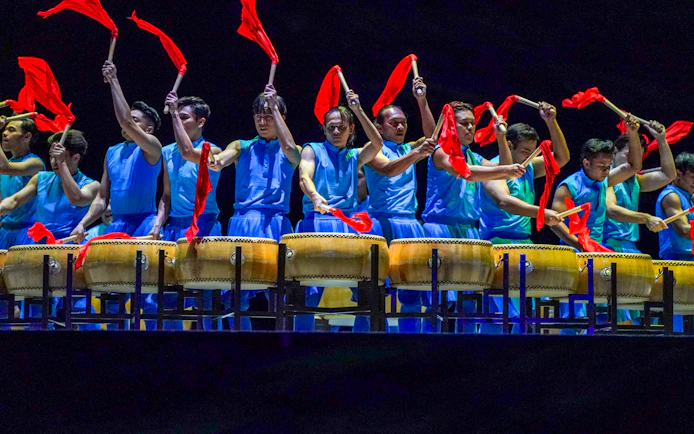 Performers in blue costumes playing drums with red flags at Encore Melaka show.