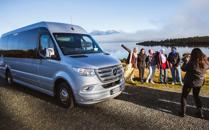 Group posing by tour van with scenic Milford Sound backdrop, Queenstown tour.