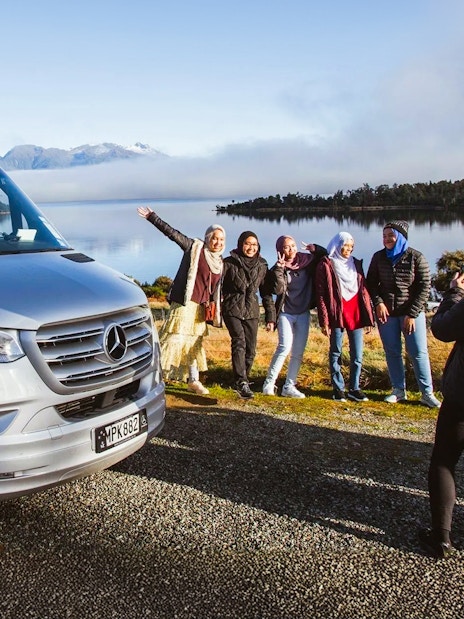 Group posing by tour van with scenic Milford Sound backdrop, Queenstown tour.