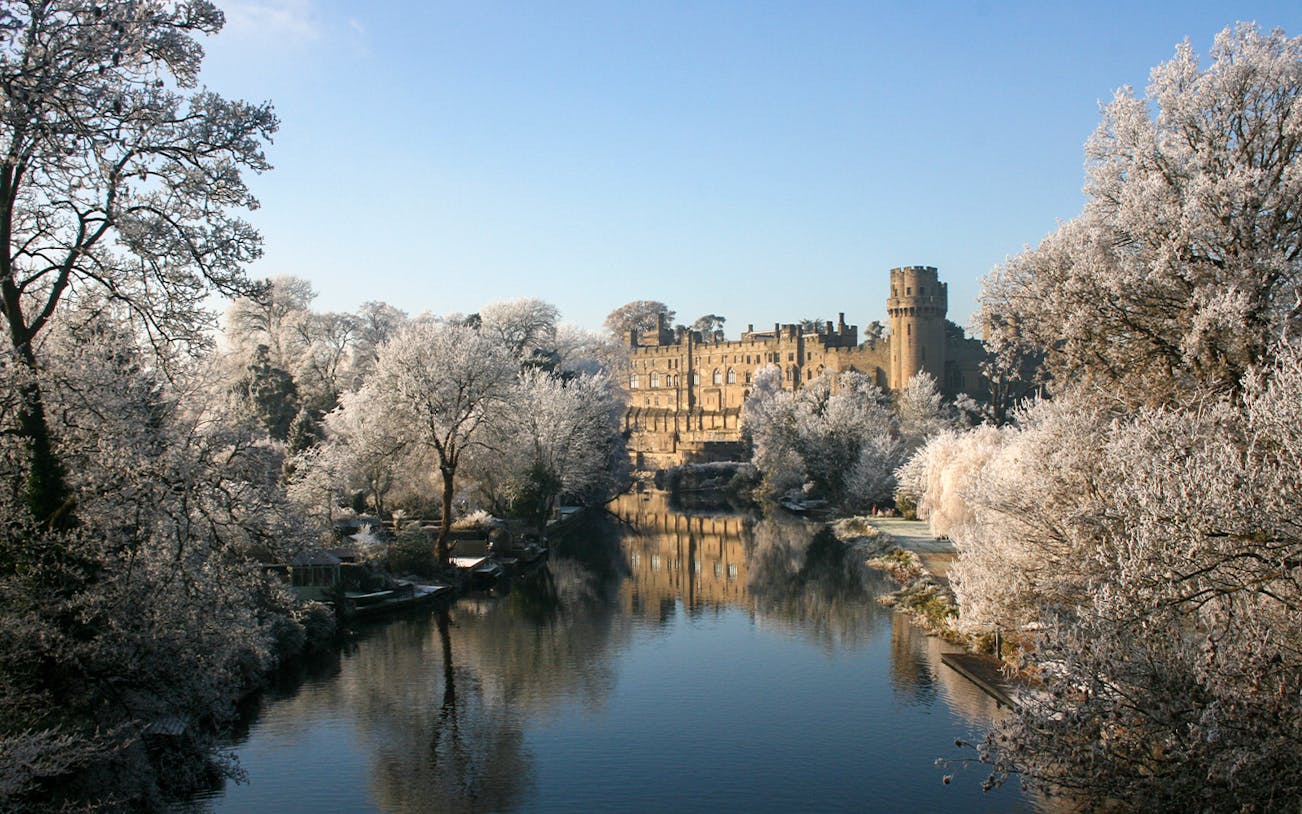 Warwick Castle in winter with frosted trees and a reflective river.