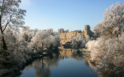 Warwick Castle in winter with frosted trees and a reflective river.