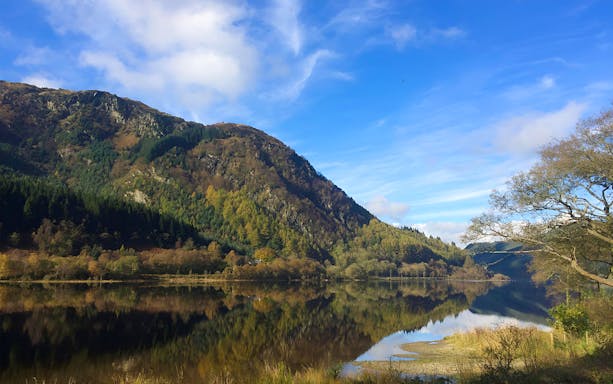 Loch Lomond with reflections of surrounding hills and trees under a blue sky.