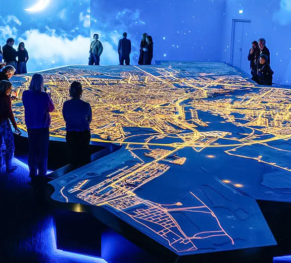 Visitors observing illuminated Amsterdam city map at the Amsterdam in Motion exhibit.