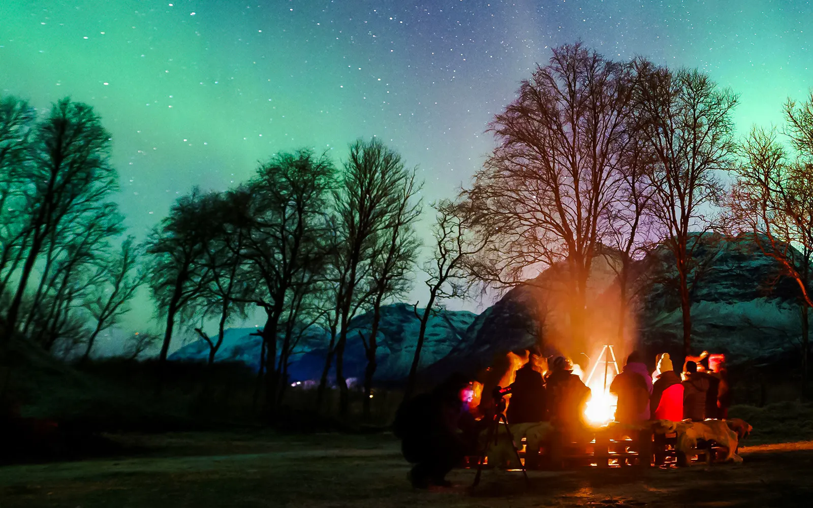 Campfire gathering under Northern Lights in Tromsø, Norway, with silhouetted trees and mountains.