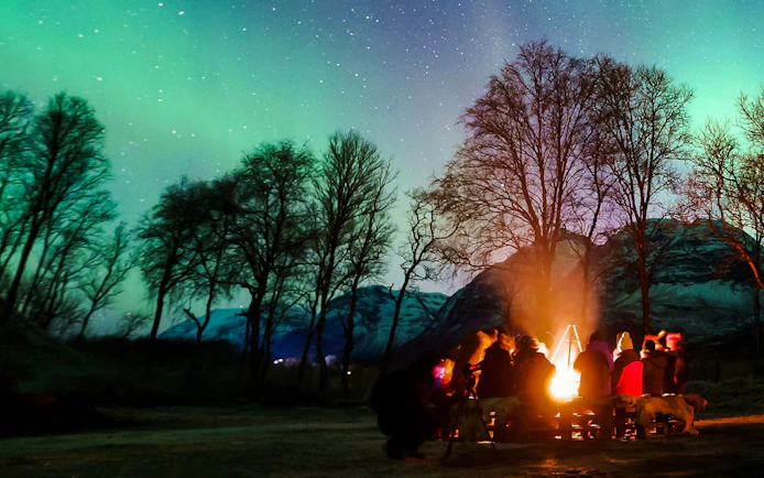 Campfire gathering under Northern Lights in Tromsø, Norway, with silhouetted trees and mountains.