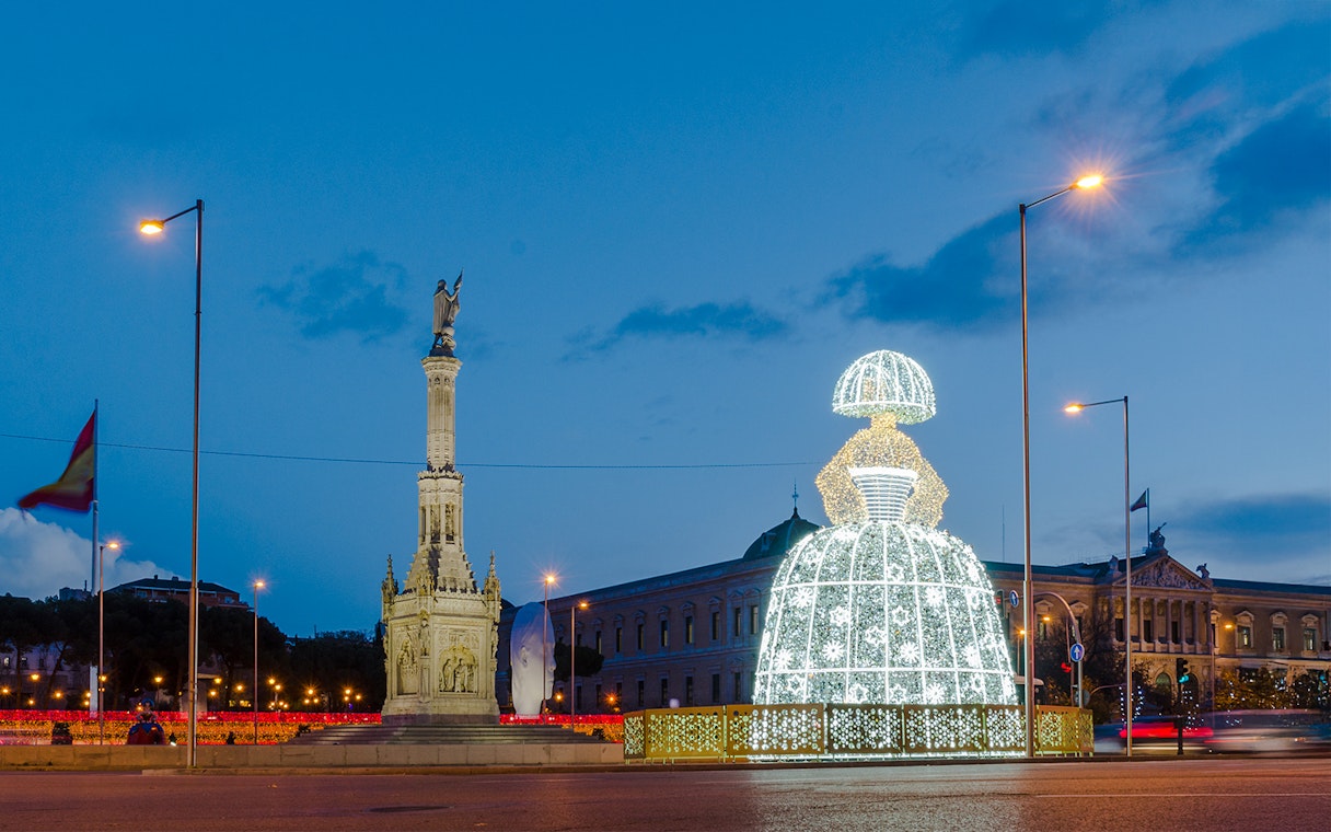 Illuminated Menina sculpture at night near a monument in Madrid, Spain during Christmas.