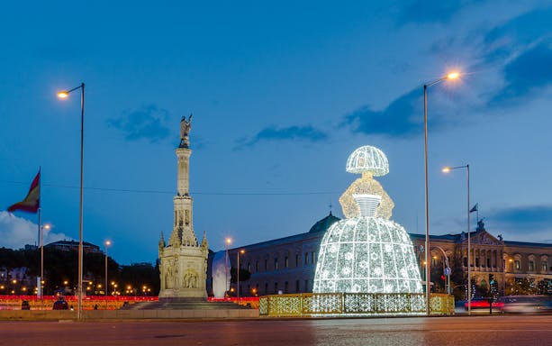 Illuminated Menina sculpture at night near a monument in Madrid, Spain during Christmas.