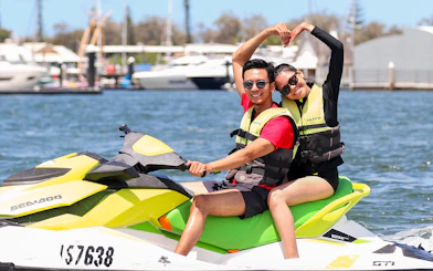 Couple enjoying a jet ski ride on a sunny day near a marina.
