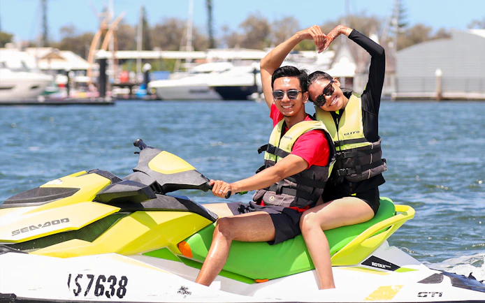 Couple enjoying a jet ski ride on a sunny day near a marina.