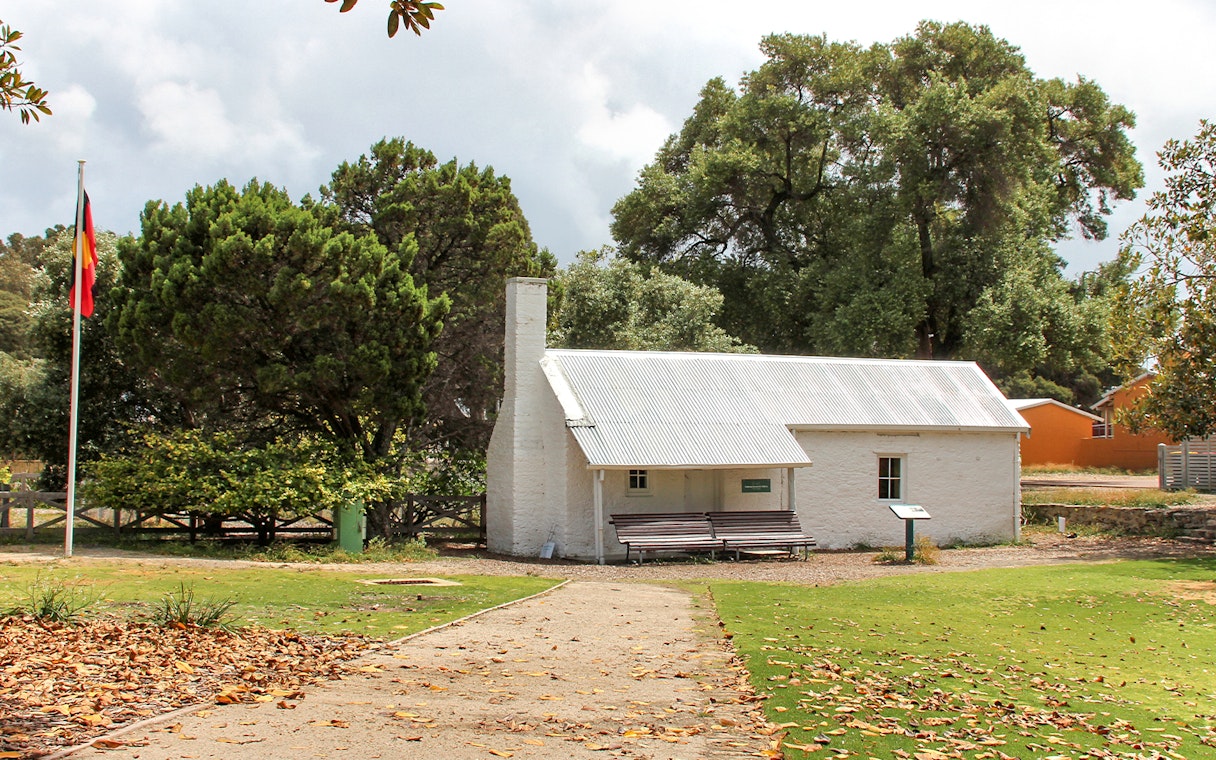 Historic cottage on Rottnest Island surrounded by trees, with a flagpole and pathway in view.