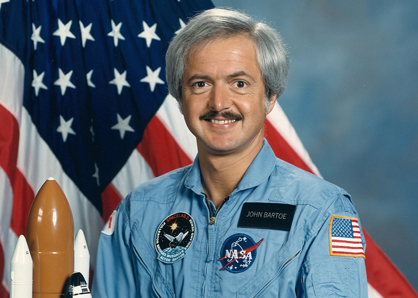 John D. Bartoe speaking at a space exploration event with a presentation screen in the background.