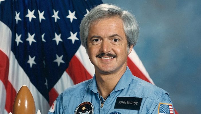 John D. Bartoe speaking at a space exploration event with a presentation screen in the background.