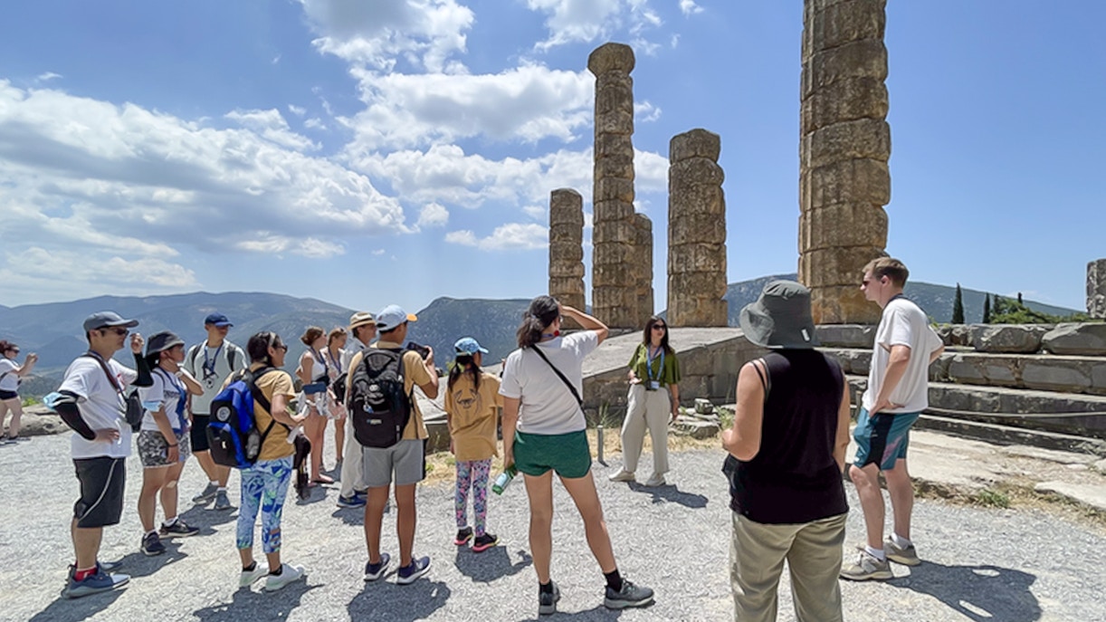 Visitors touring ancient ruins at Delphi, Greece, with guide explaining historical significance.
