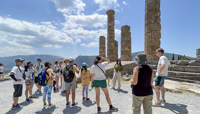 Visitors touring ancient ruins at Delphi, Greece, with guide explaining historical significance.