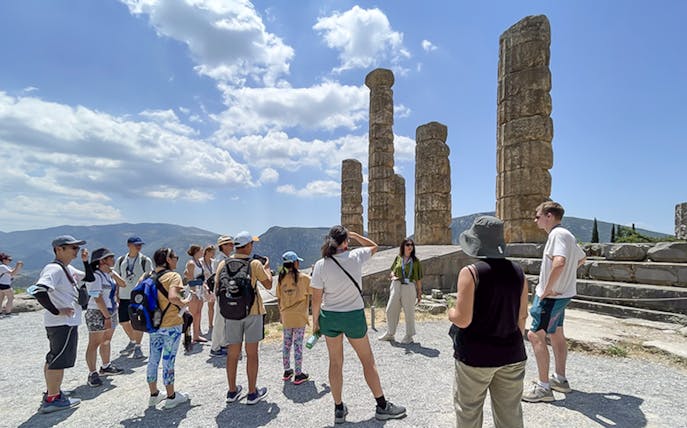 Visitors touring ancient ruins at Delphi, Greece, with guide explaining historical significance.