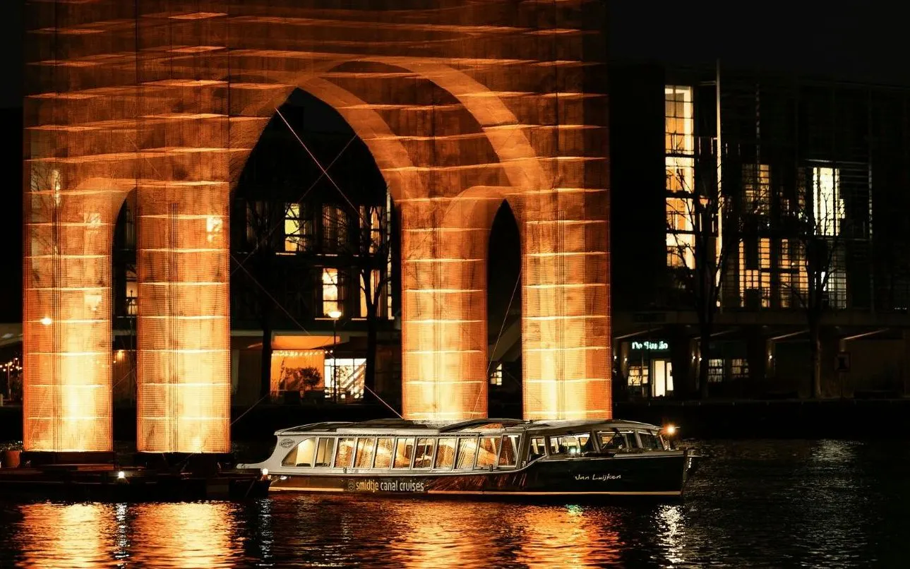 Canal cruise passing illuminated arch at Amsterdam Light Festival.