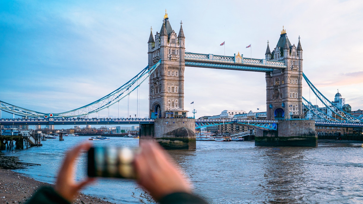 Tower Bridge in London with a person taking a photo, view from the riverbank.