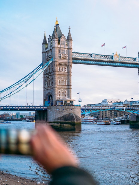 Tower Bridge in London with a person taking a photo, view from the riverbank.