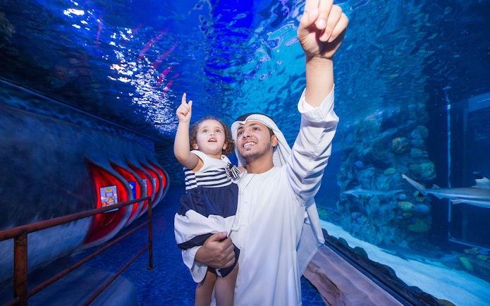 Father and child pointing at marine life in Dubai Aquarium tunnel.
