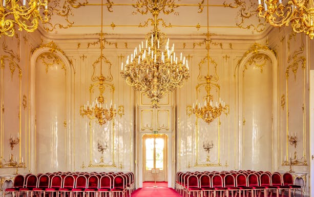 Royal Palace of Gödöllő interior with ornate chandeliers and red velvet chairs.