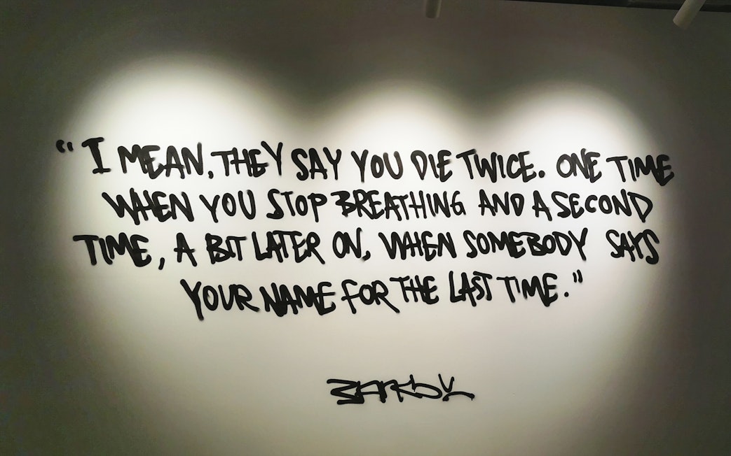 Banksy artwork with quote about dying twice, displayed on a wall.