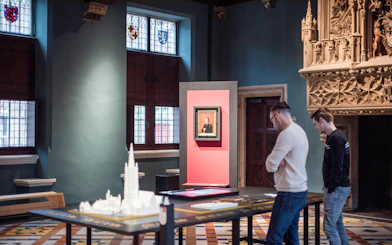 Visitors observing exhibits at Gruuthusemuseum, Bruges, with historical artifacts and stained glass windows.