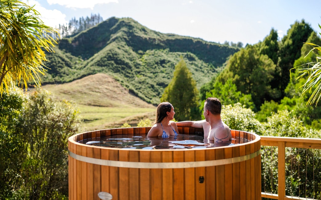 Couple relaxing in a hot tub with scenic view of lush hills at Waitomo, New Zealand.