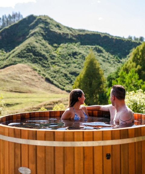 Couple relaxing in a hot tub with scenic view of lush hills at Waitomo, New Zealand.