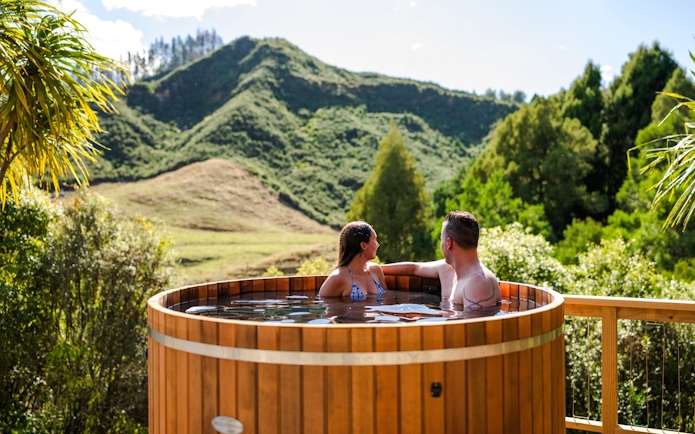 Couple relaxing in a hot tub with scenic view of lush hills at Waitomo, New Zealand.