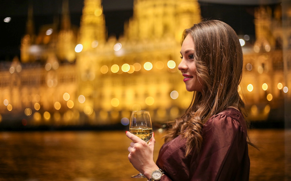 Woman enjoying a drink on Budapest dinner cruise with illuminated cityscape.