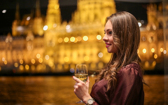 Woman enjoying a drink on Budapest dinner cruise with illuminated cityscape.