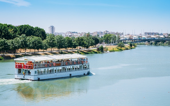 Cruise boat on Guadalquivir River in Sevilla with cityscape and trees in the background.
