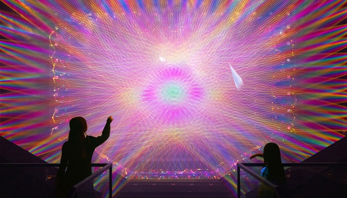 Two figures observe a vibrant digital light installation with colorful patterns.