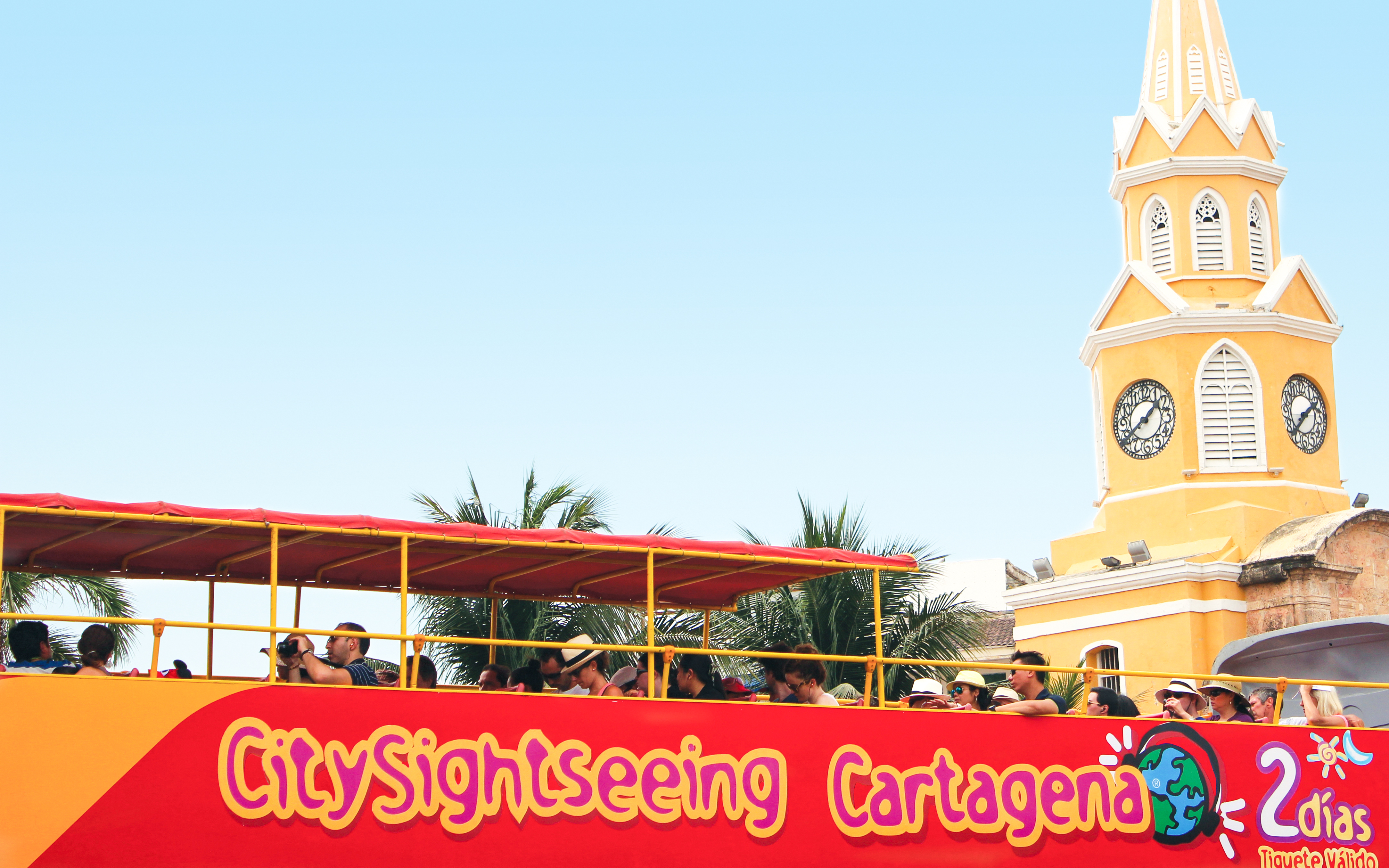 People on upper deck of City Sightseeing bus in Cartagena, near historic clock tower.