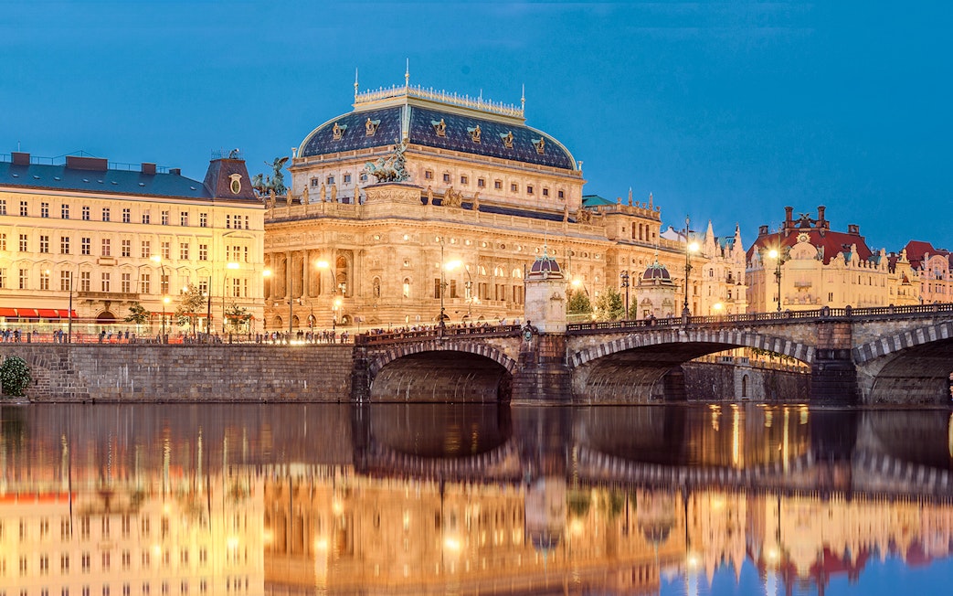 Prague's National Theatre and bridge illuminated at night, viewed from a river.