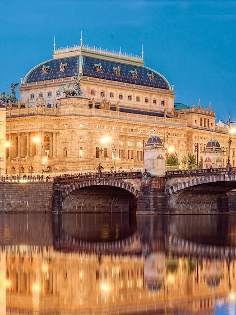 Prague's National Theatre and bridge illuminated at night, viewed from a river.
